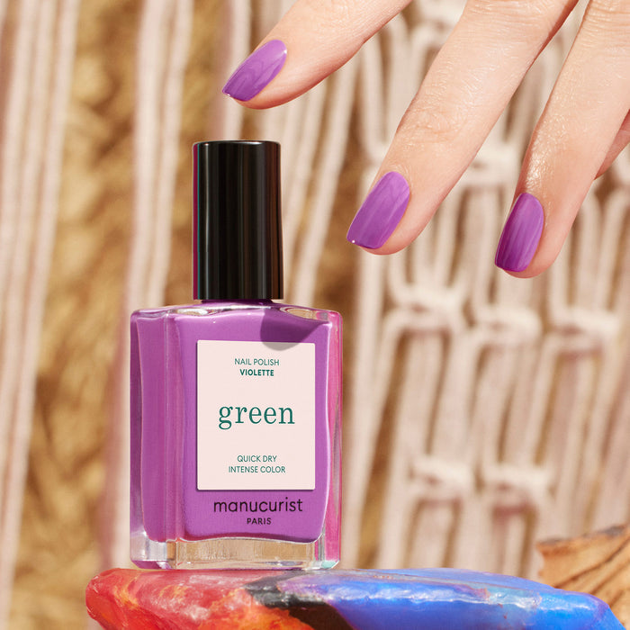 A hand with retro purple painted nails hovers above a Violette nail polish bottle by Manucurist, set on a colorful surface with a woven background.
