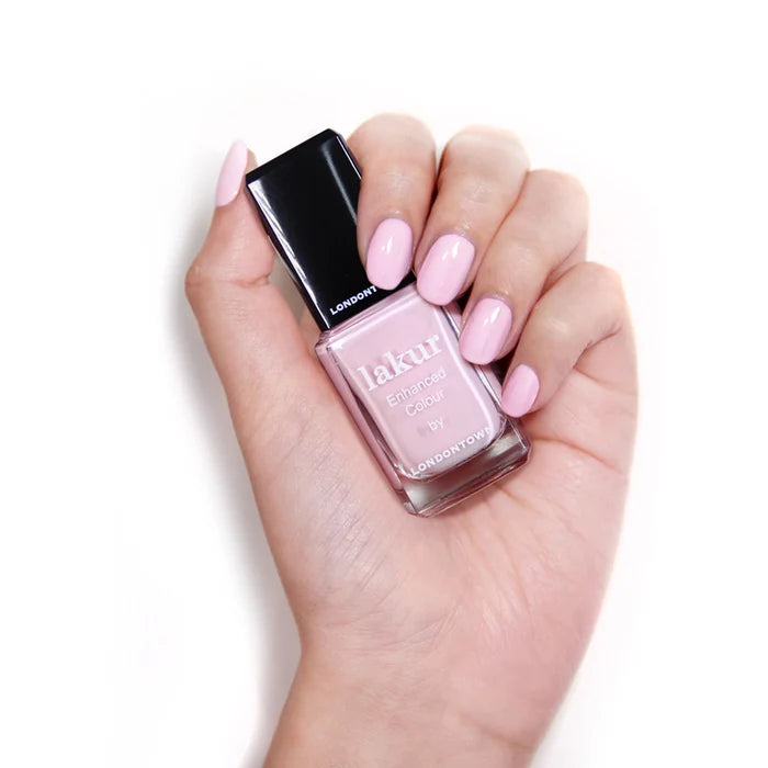 A hand with light pink painted nails holds a bottle of LONDONTOWN's Afternoon Tea nail polish in a matching shade against a white background.