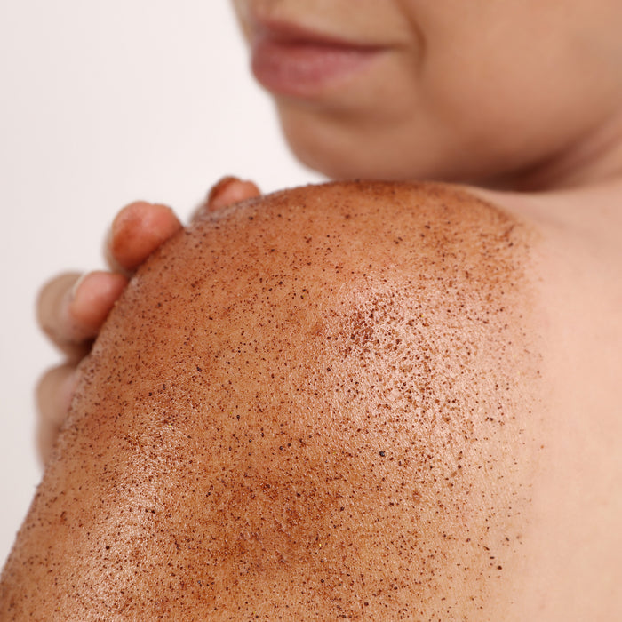 Close-up of exfoliating body scrub with fine granules applied to shoulder skin, showing gentle texture for all skin types.