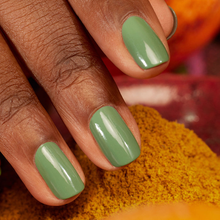 A close-up of a hand with medium brown skin, nails polished in Manucurist's glossy "Artichoke" shade. Warm yellow and orange tones in the background evoke a Moroccan garden atmosphere.