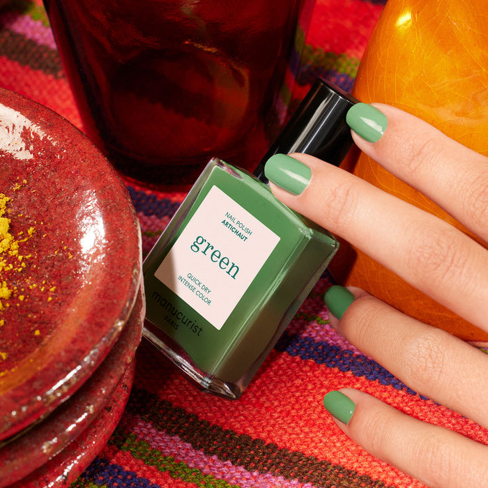 A hand with green-painted nails holds a Manucurist nail polish bottle labeled Artichoke, resting on vibrant woven fabric and surrounded by red and orange ceramics for a Moroccan garden vibe.