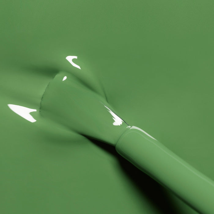 A close-up of a brush applying Manucurist’s Artichoke nail polish to a smooth surface, with light reflecting off the glossy, herbal green paint.