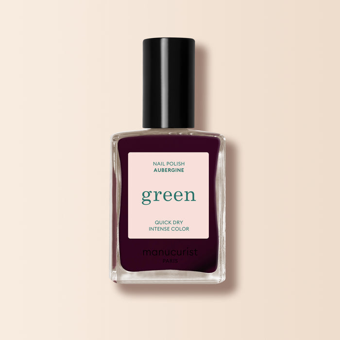 A bottle of Manucurist Aubergine nail polish, with a deep purple shade and a black cap, stands against a light beige background.