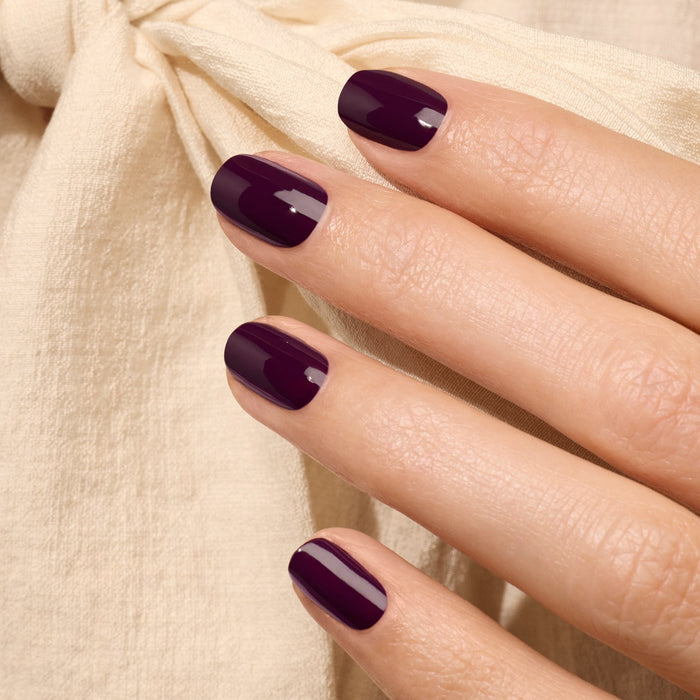 A hand with neatly manicured nails painted in Aubergine by Manucurist rests on light beige fabric featuring a soft, tied bow.
