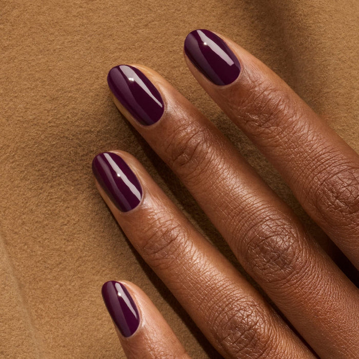 A hand with glossy, dark nails—painted in the deep purple "Aubergine" shade from Manucurist—rests on a soft, light brown textured surface.