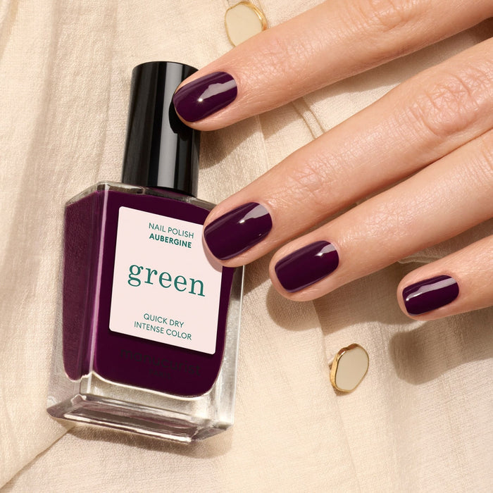 A hand with neatly manicured nails painted deep purple holds a bottle of Manucurist Aubergine nail polish, resting on cream fabric with gold accents.