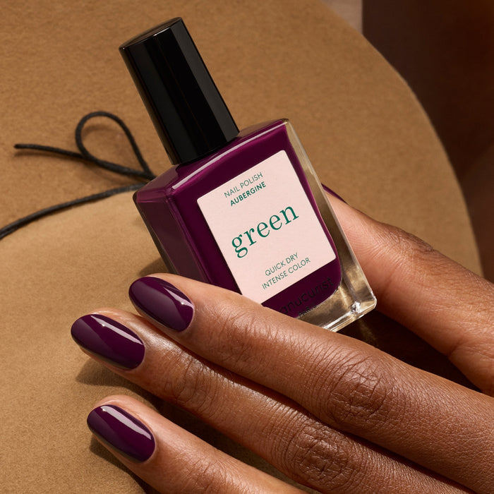 A hand with dark purple nail polish holds a matching Manucurist bottle labeled “Aubergine” from the Green™ range, showing “QUICK DRY INTENSE COLOR,” set against a tan background.