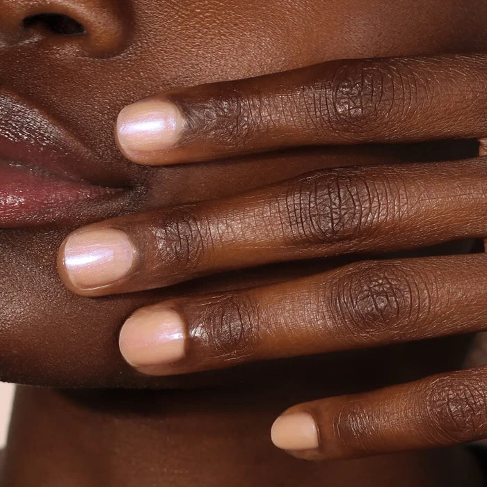 A close-up of a hand with neatly manicured, short natural nails painted in LONDONTOWN's Bare Illuminating Nail Concealer, a pale pink shimmery polish, held near the person's face.