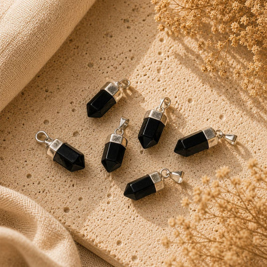 Five black onyx crystal point pendants with silver-toned caps and loops, arranged on a textured beige stone surface with dried flowers and beige fabric.