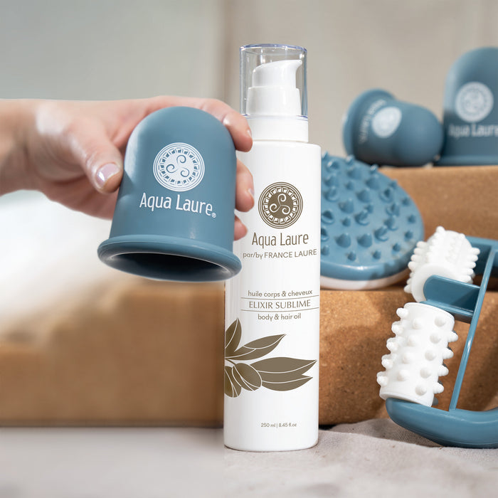 Aqua Laure Elixir Sublime body & hair oil bottle with pump, held beside a blue silicone applicator, surrounded by massage tools for skincare routine.