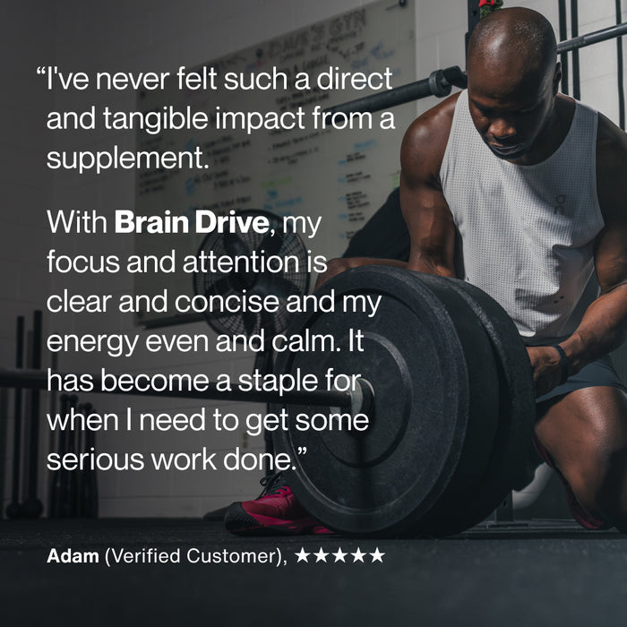 A man in a gym kneels beside a loaded barbell while wearing a white tank top and red shoes. Text on the image reads: “I’ve never felt such a direct and tangible impact from a supplement. With Brain Drive, my focus and attention is clear and concise and my energy even and calm. It has become a staple for when I need to get some serious work done.” “Adam (Verified Customer), ★★★★★”