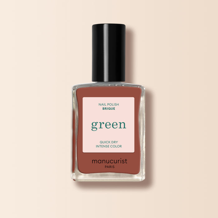 A bottle of Manucurist Brique nail polish from the Green™ range, featuring a brick red shade with a black cap and label. The rich color is inspired by Moroccan street markets, set against a light beige background.
