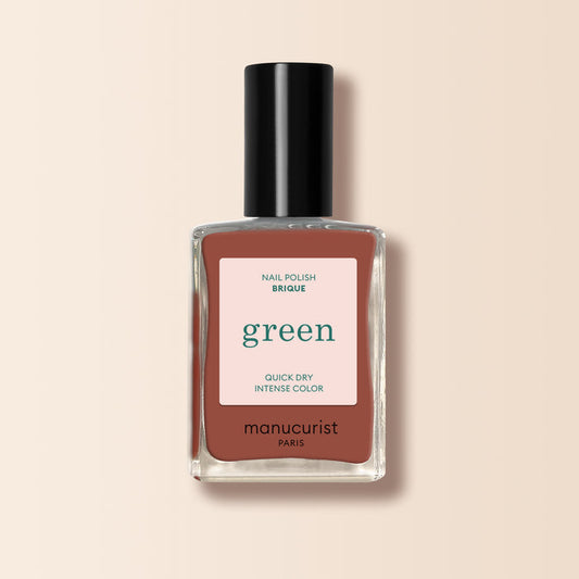 A bottle of Manucurist Brique nail polish from the Green™ range, featuring a brick red shade with a black cap and label. The rich color is inspired by Moroccan street markets, set against a light beige background.
