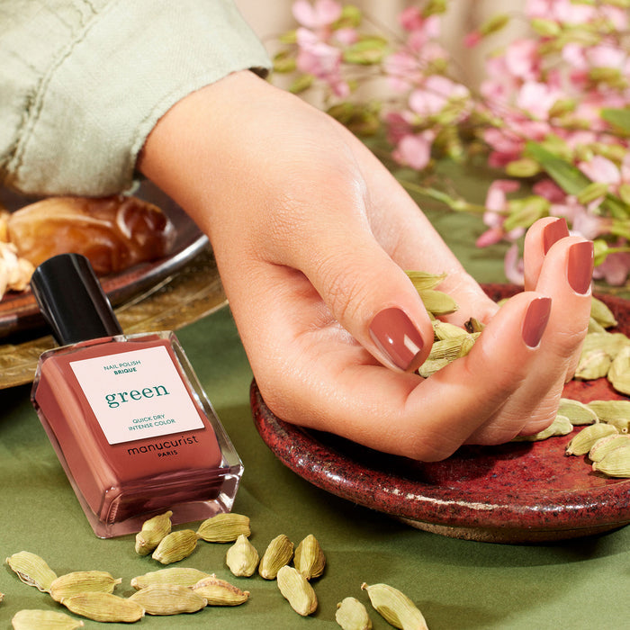 A hand with glossy, brownish-pink nails painted in Manucurist's Brique from the Green™ range rests on a red plate with cardamom pods, evoking a Moroccan market. A matching Brique bottle sits beside it, with blossoms and leaves in the background.