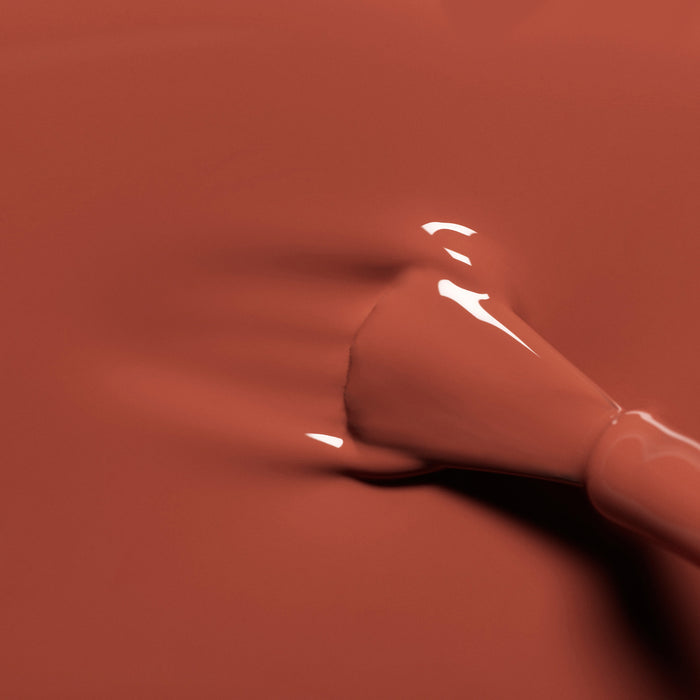 A close-up of a Manucurist Brique brush spreading glossy, brick red nail polish on a smooth surface, with reflections highlighting the wet finish.