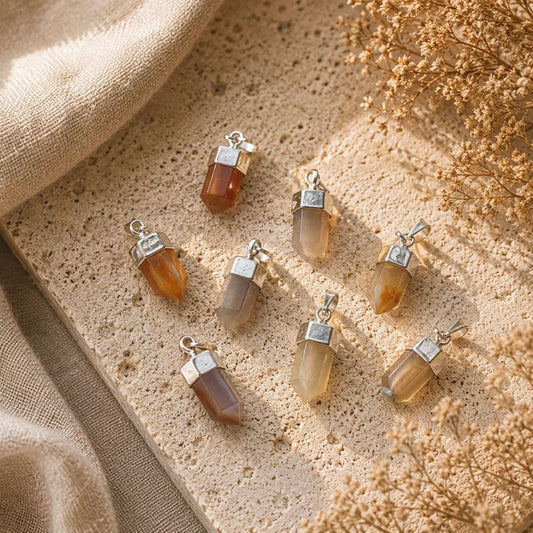 Six small pendant charms with translucent stone drops and silver caps, arranged on a textured beige surface with linen and dried flowers.