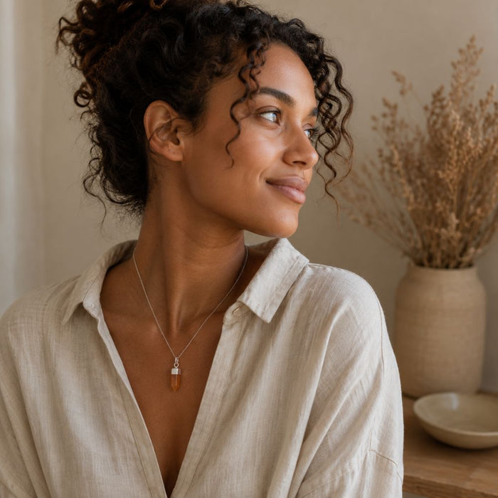 A woman with curly dark hair wearing a silver-toned pendant necklace with a small amber-orange stone, paired with a light beige linen shirt. She is shown in a warm, softly lit indoor setting with dried flowers in a vase in the background.