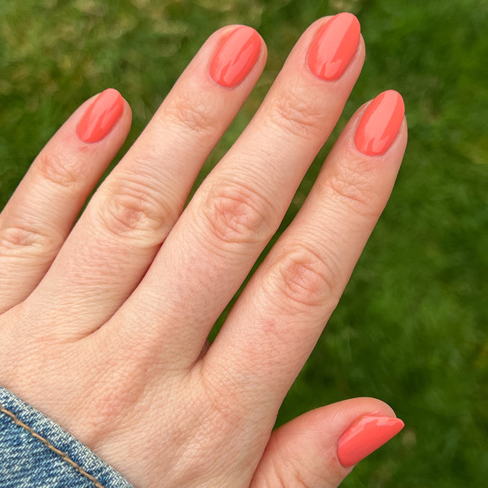 Hand with oval nails painted bright coral in Carrot Juice gel polish by Olive and June against green grass background