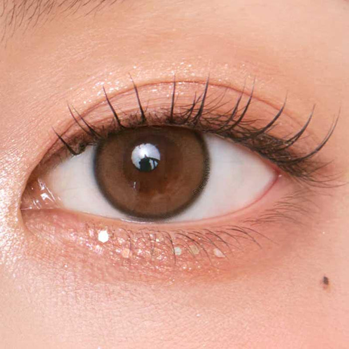 A close-up of a brown eye with long lashes, soft peach eyeshadow, and smooth skin, enhanced naturally by Chuu Aery Rose Lucid Brown (10pk) color contacts from Chuu. A beauty mark sits below the lower lash line.