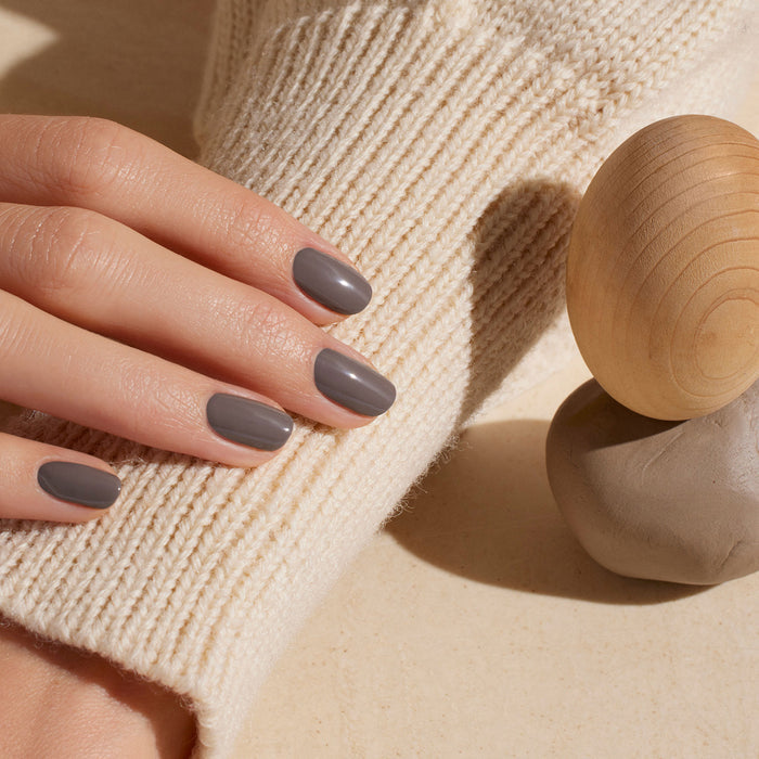 A hand with short, neatly manicured nails painted in Manucurist Clay rests on a cream-colored knitted sweater beside a wooden sphere and a clay object.