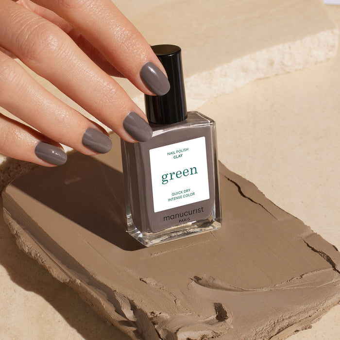 A hand with short, neat nails painted in Clay by Manucurist holds the matching nail polish bottle, set against neutral clay-toned surroundings—elegantly showcasing this timeless taupe shade.