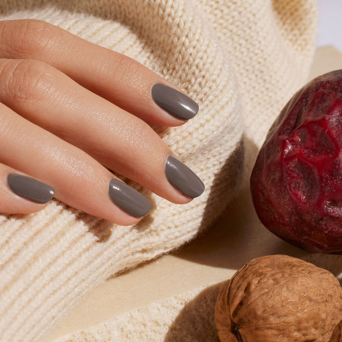 A hand with neatly manicured nails painted in Manucurist’s Clay shade rests on a cream-colored knitted sweater, next to a walnut and a red pomegranate.