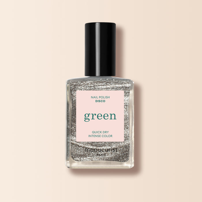 A clear glass bottle of Manucurist Disco silver glitter nail polish from the Green™ range, labeled green and quick dry intense color, stands upright against a light beige background.
