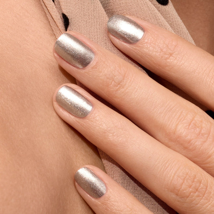 A close-up of a hand with short, neatly manicured nails painted in Manucurist’s Disco, an opaque glittery silver polish, resting on beige fabric with black dots.