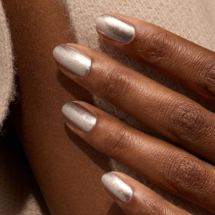 A close-up of a hand with medium-brown skin, nails neatly manicured and painted with Manucurist’s Disco—an opaque glittery silver nail polish—resting on soft beige fabric.