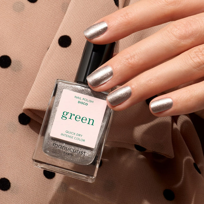 A hand with opaque glittery silver nail polish holds a bottle of Manucurist Disco by Manucurist, set against beige fabric with black polka dots.