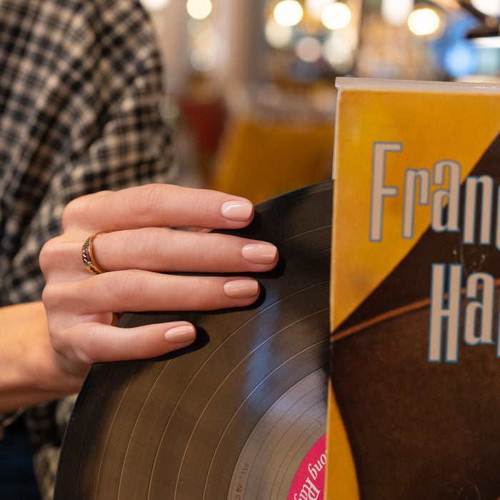 A person with neatly manicured nails in Manucurist’s Dune holds a vinyl record near a yellow-orange album cover partially showing the text "Fran.