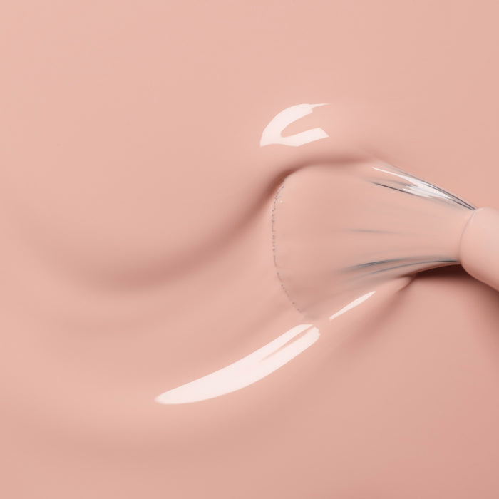 A close-up of a brush swirling Manucurist Dune foundation, a creamy light pink shade with orange undertones, on a smooth surface, creating a glossy texture and softly blurred streaks.