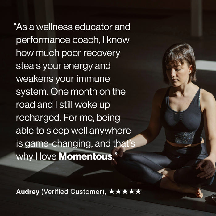Woman sitting cross-legged indoors in a dimly lit room. Text reads: “As a wellness educator and performance coach, I know how much poor recovery steals your energy and weakens your immune system. One month on the road and I still woke up recharged. For me, being able to sleep well anywhere is game-changing, and that’s why I love Momentous.” Attribution reads: “Audrey (Verified Customer), ★★★★★.”
