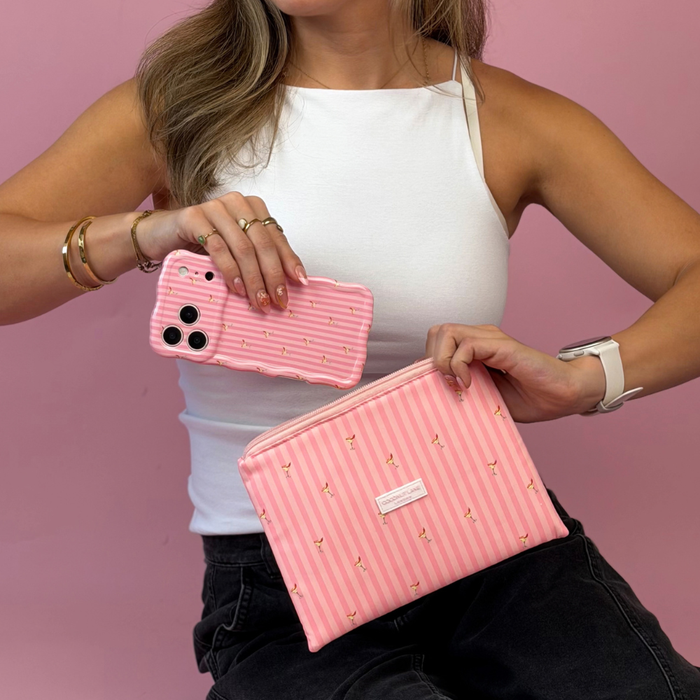 A woman holds a pink striped phone case in one hand and a pink zippered cosmetic pouch with vertical pink stripes and small spicy margarita illustration prints on it.