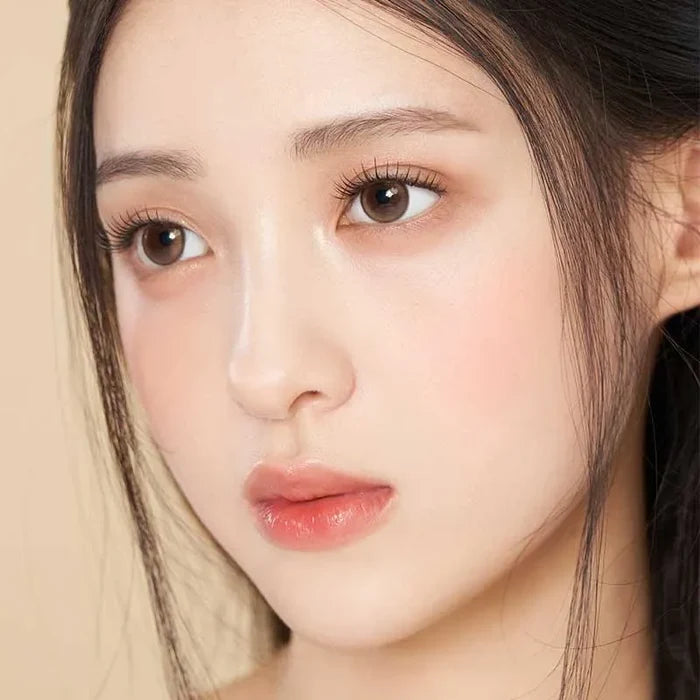 A close-up of a young woman with fair skin and straight dark hair wearing Gemhour Demeter 1-Day 13.5 Amber Brown contacts (10pk), which highlight her brown eyes, long lashes, and glossy pink lips as she gazes thoughtfully to the side.
