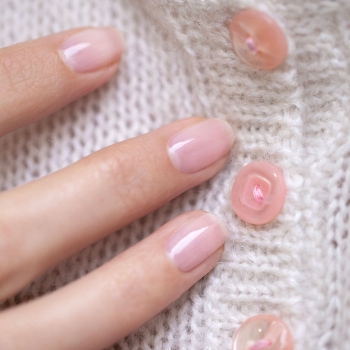 A hand with neatly manicured nails painted in Hortencia by Manucurist, a sheer pink polish, rests on soft white knitted fabric decorated with pink buttons.