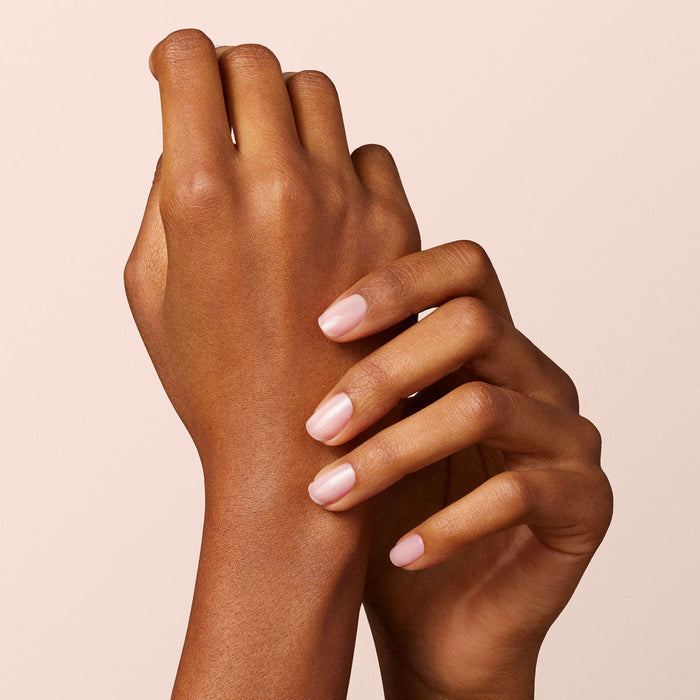 Two hands with smooth brown skin are seen against a neutral background. One gently holds the other's wrist, showing neatly manicured nails painted with Manucurist's Hortencia—a sheer pink nail polish.