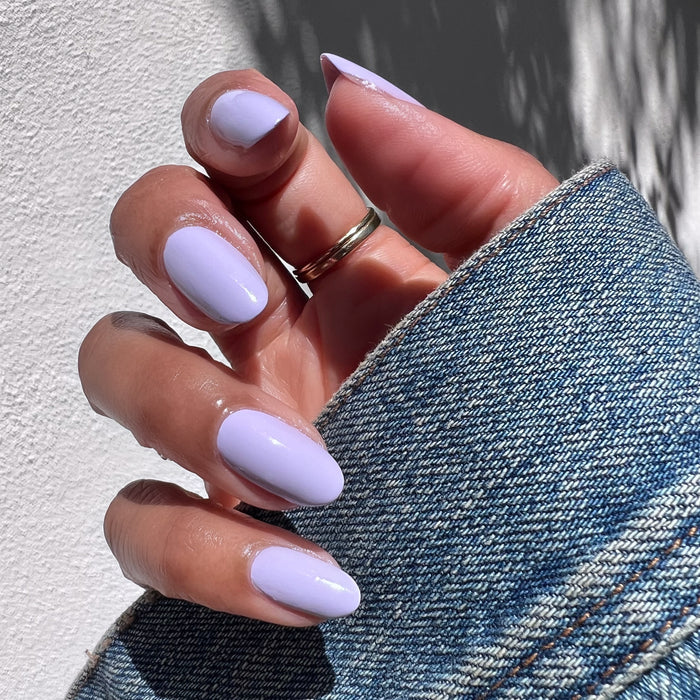 Hand with almond-shaped nails painted in pastel lavender Jacaranda gel polish against a white background with denim sleeve