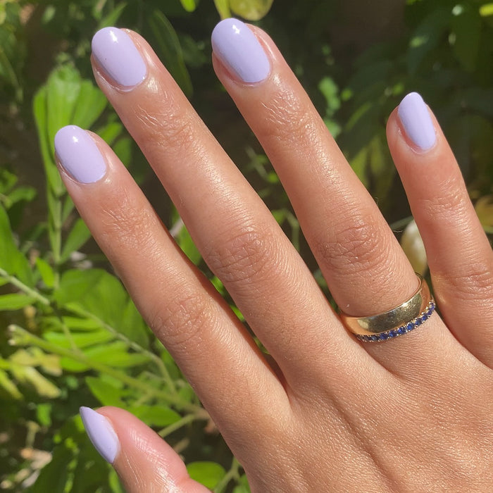 Hand with nails painted in pastel lavender Jacaranda quick dry gel polish outdoors with gold rings and greenery background