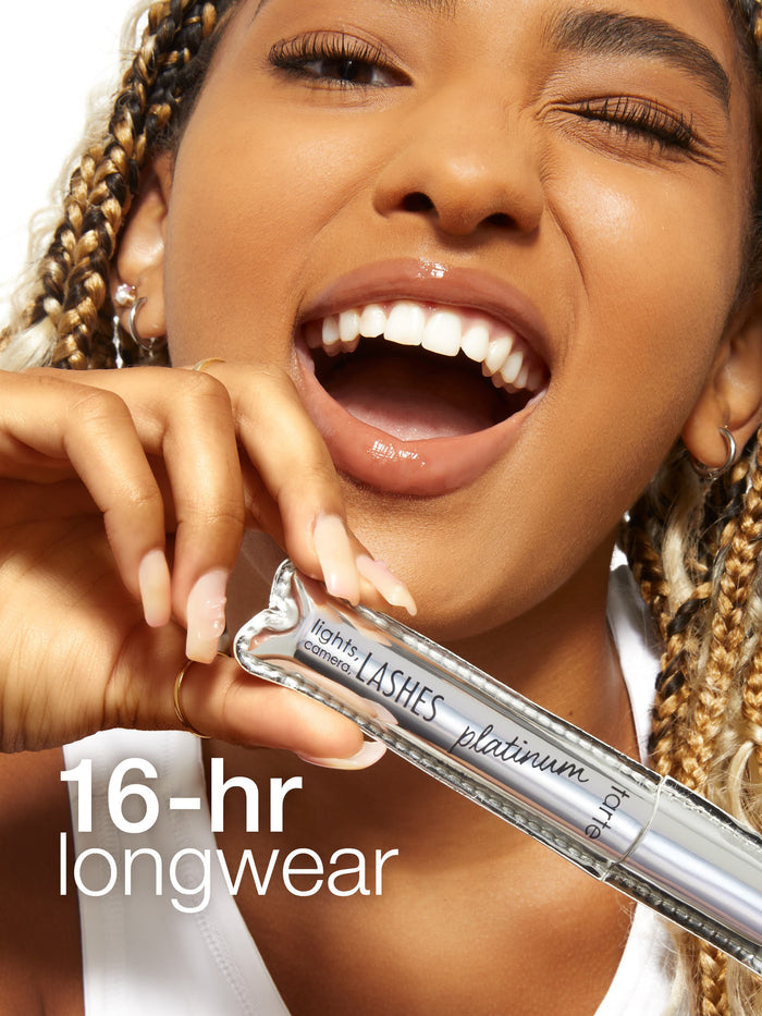 A person joyfully holds Tarte Cosmetics' lights, camera, LASHES platinum mascara in front of their face. The text reads, 16-hr longwear. The background is a plain white surface.