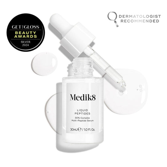 A clear glass dropper bottle of Liquid Peptides serum is presented, with its dropper removed and resting beside it and serum droplets, showing the texture.