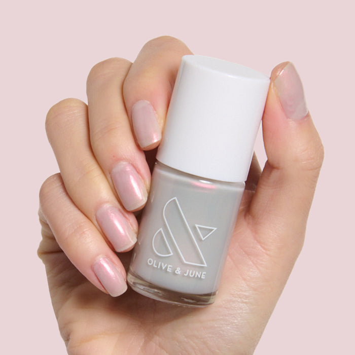 Hand holding Olive and June Lucky Oyster bottle with nails painted in shimmery grey iridescent polish for a soft manicure