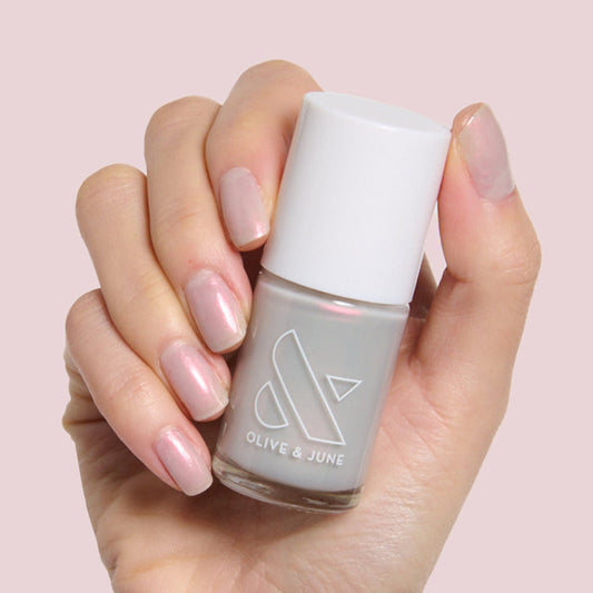 Hand holding Olive and June Lucky Oyster bottle with nails painted in shimmery grey iridescent polish for a soft manicure