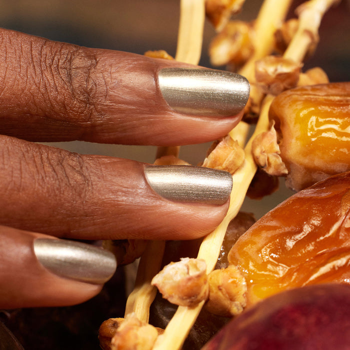 Fingers with Manucurist Metallic nail polish in a cool-toned silver shade touch a cluster of dried dates on their stem.