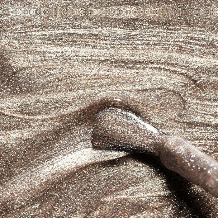 A close-up of the Manucurist Metallic makeup brush swirling through shimmery bronze powder, creating glittery textured streaks with cool-toned metallic highlights.