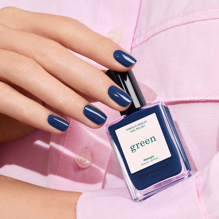 A hand with neatly manicured nails painted Midnight holds a matching Manucurist bottle, set against a light pink shirt background.