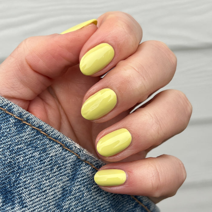 Hand with short oval nails painted vibrant yellow-green gel polish in Not Kale by Olive and June against denim background