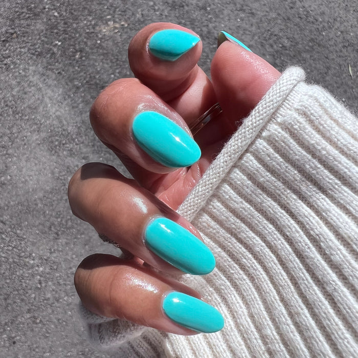Hand with nails painted glossy electric turquoise Parakeet gel polish shown in natural light with cream sweater sleeve