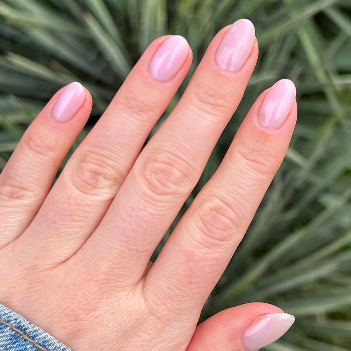 Hand with nails coated in Pink Capiz showing a glossy milky pink manicure against a green plant background