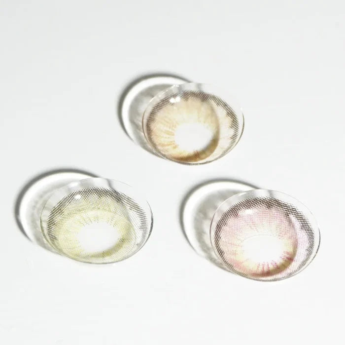 Three EyeCandys circle lenses in subtle brown, green, and pinkish shades are arranged on a white surface with soft shadows. The EyeCandys Pink Label Angel Brown lens stands out for its natural, elegant tint.